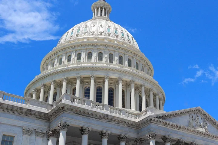White domed government building with ornate columns and architectural details viewed from below against a bright blue sky with scattered clouds.