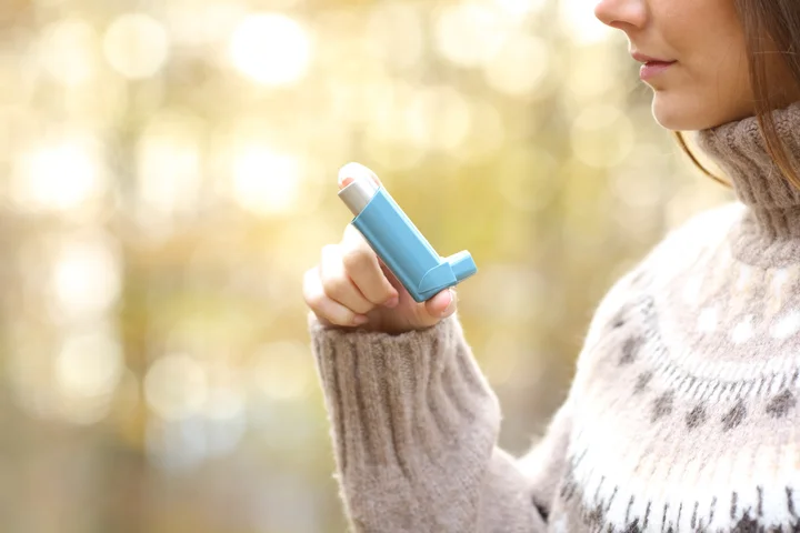 Person holding a blue inhaler near the mouth, outdoors with a blurred background.