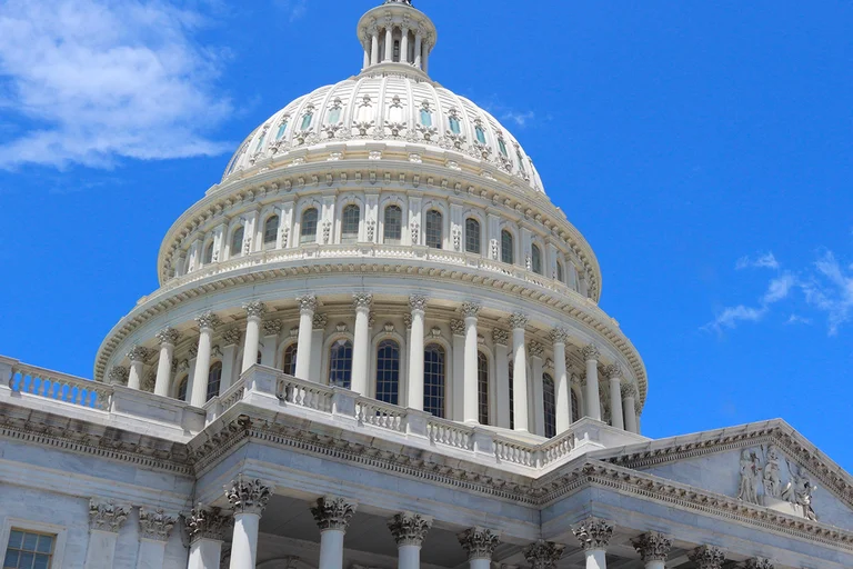 White domed government building with ornate columns and architectural details viewed from below against a bright blue sky with scattered clouds.