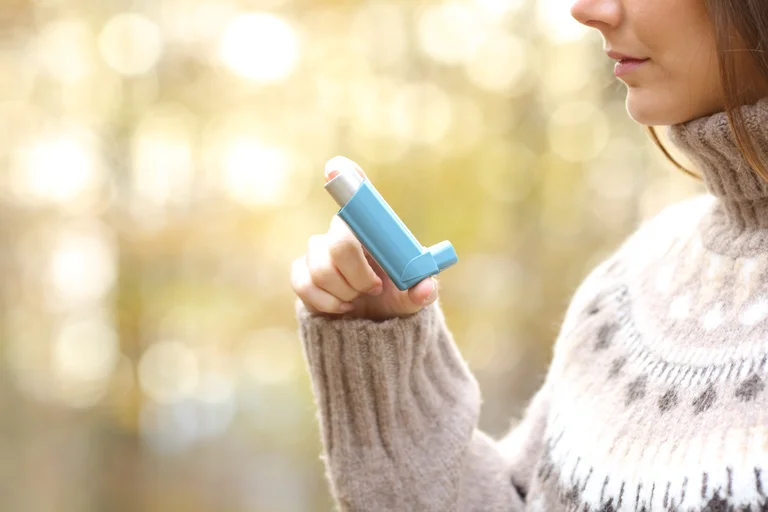 Person holding a blue inhaler near the mouth, outdoors with a blurred background.
