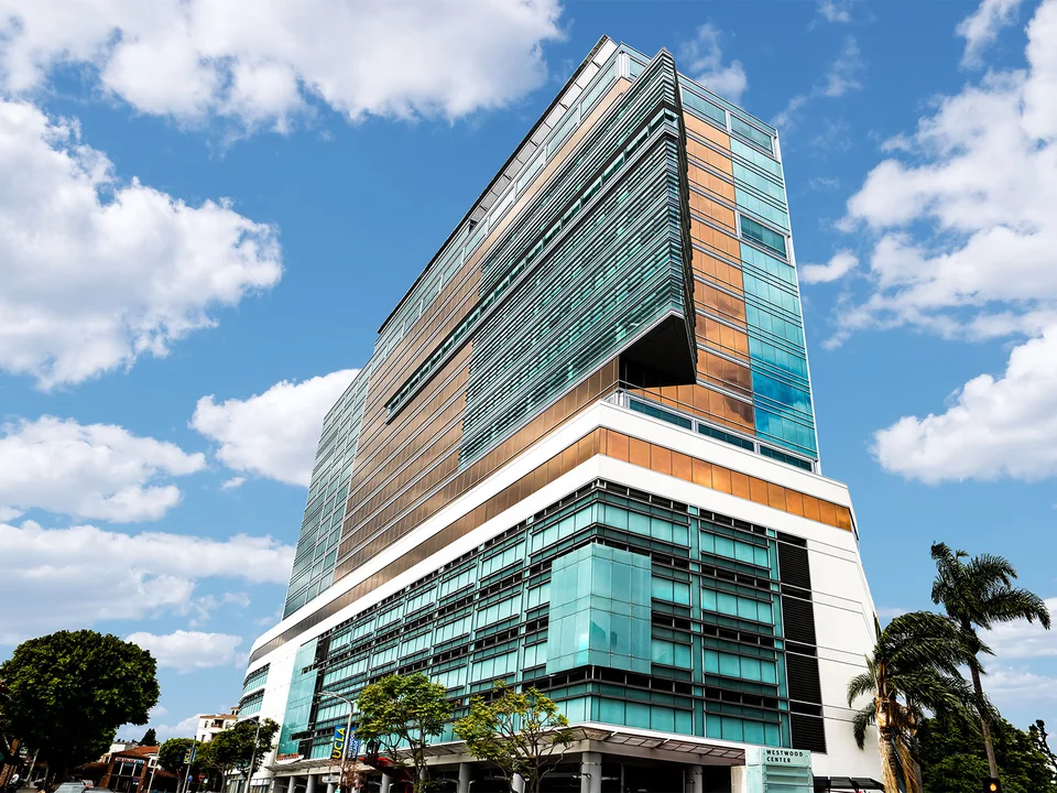 Modern multi-story glass and metal building with teal accents, viewed looking up against a partly cloudy sky.