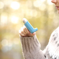 Person holding a blue inhaler near the mouth, outdoors with a blurred background.